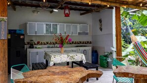 An outdoor dining area with a wooden table and chairs, a kitchen in the background, and a hammock. - HOSTAL VILLA GUADUA TAYRONA (Santa Marta)