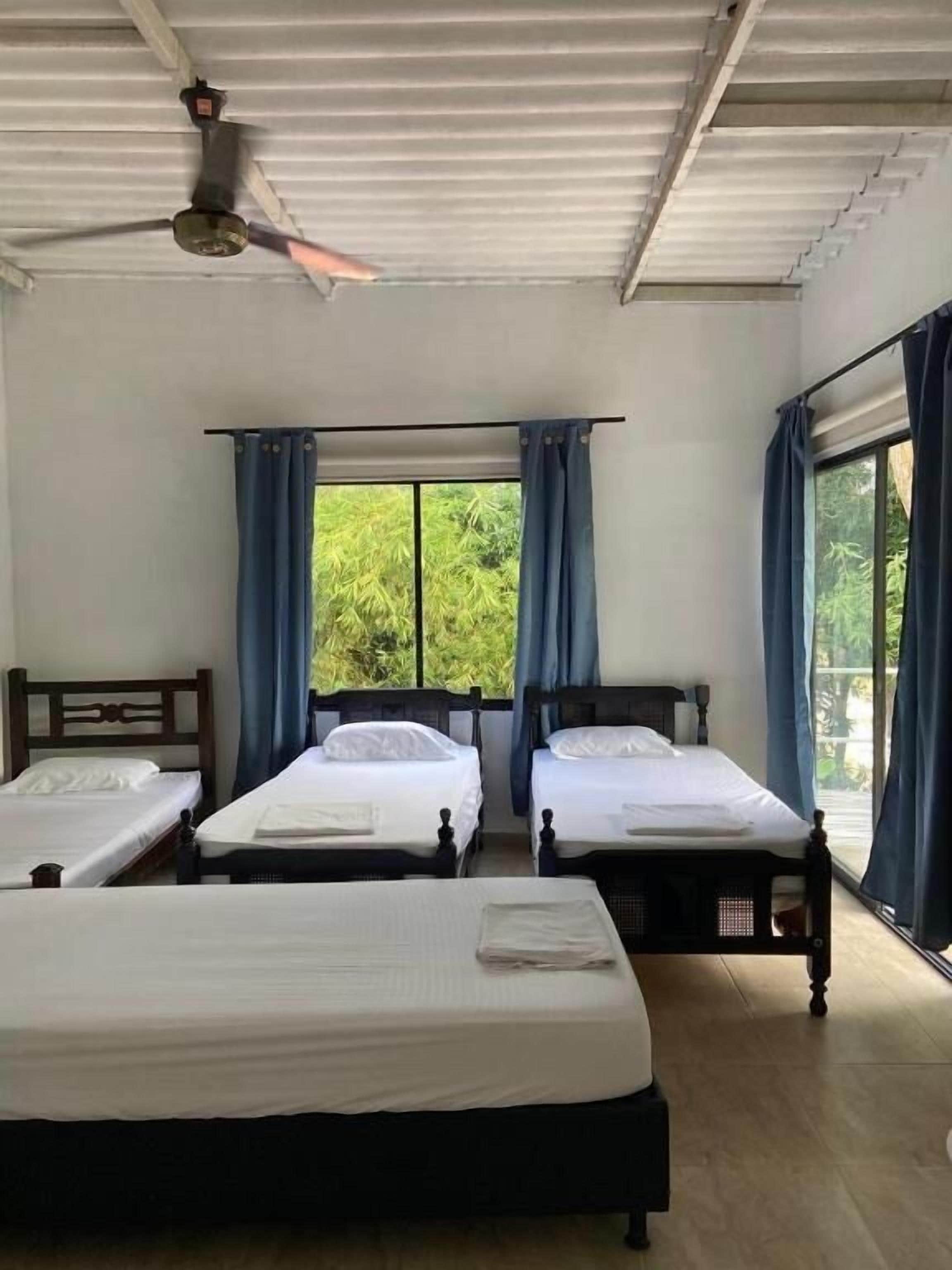Quarto com três camas de solteiro, ventilador de teto e janelas amplas com cortinas.