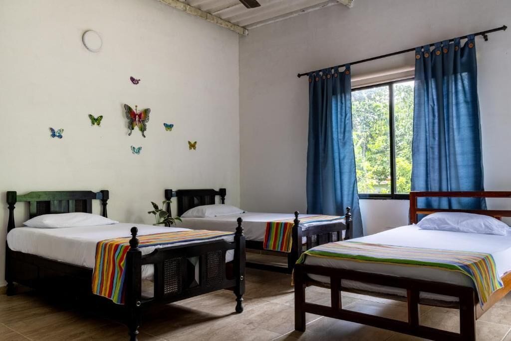 Une pièce avec trois lits en bois, chacun doté d’un matelas blanc et de draps à rayures. On y trouve des autocollants décoratifs représentant des papillons sur les murs, une fenêtre avec des rideaux bleus et un ventilateur de plafond.
