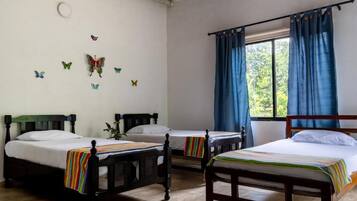 Une pièce avec trois lits en bois, chacun doté d’un matelas blanc et de draps à rayures. On y trouve des autocollants décoratifs représentant des papillons sur les murs, une fenêtre avec des rideaux bleus et un ventilateur de plafond.