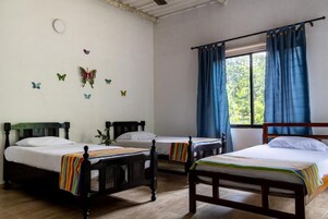 Una habitación con tres camas de madera, cada una con colchón blanco y ropa de cama a rayas. Hay adhesivos de mariposas en la pared, una ventana con cortinas azules y un ventilador de techo.