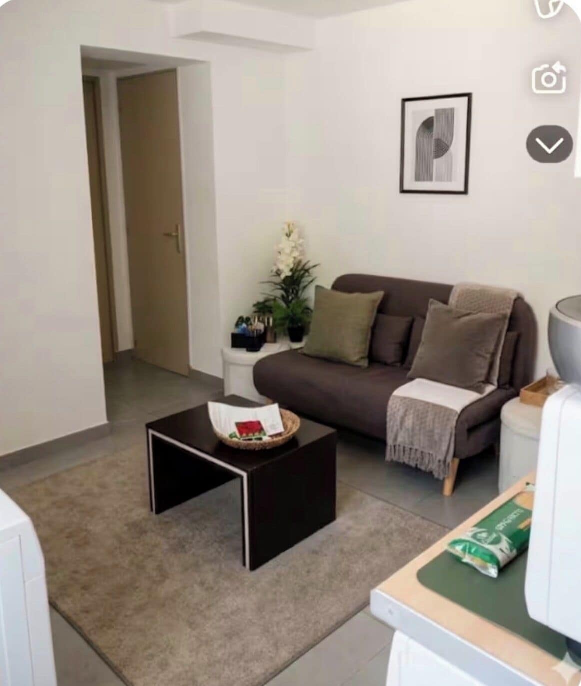 A living room with a brown sofa, a black coffee table, a white side table, and a framed picture on the wall.