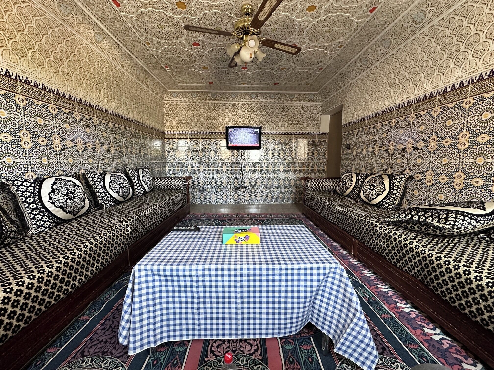 A room with a checkered table, patterned sofas, a ceiling fan, and a television.