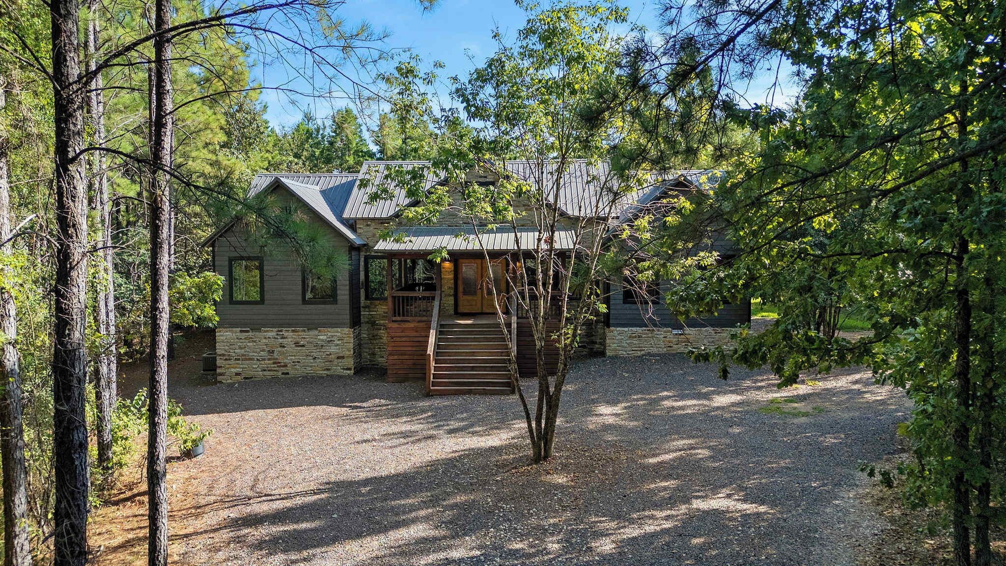 A cabin with a stone foundation, wooden exterior, and a metal roof, surrounded by trees and gravel.