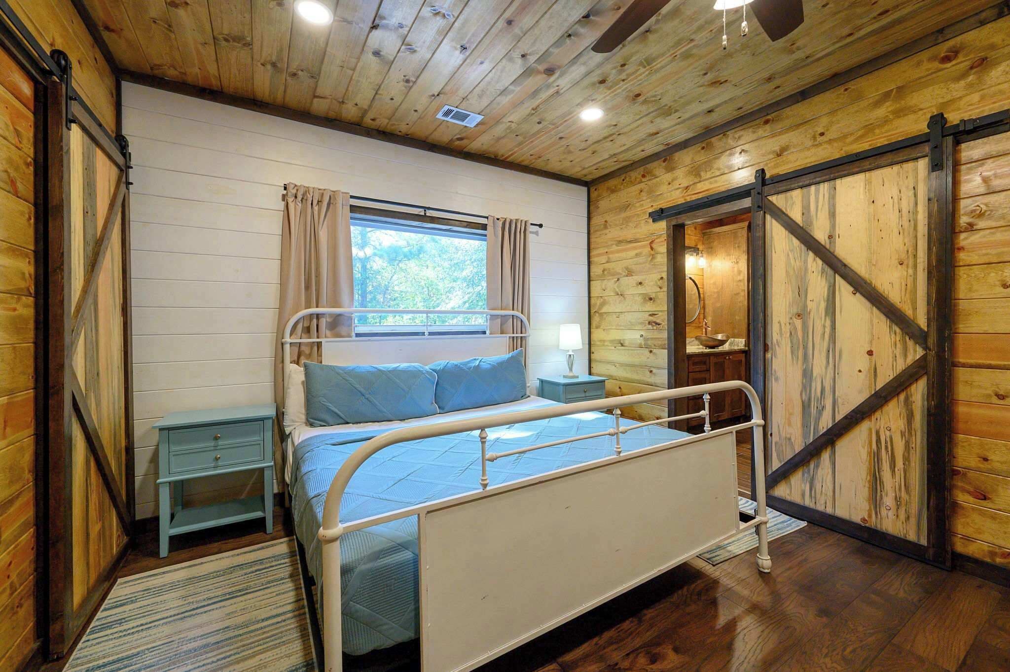 A wooden cabin bedroom with a metal bed frame, a window with curtains, a nightstand, and a wooden door.