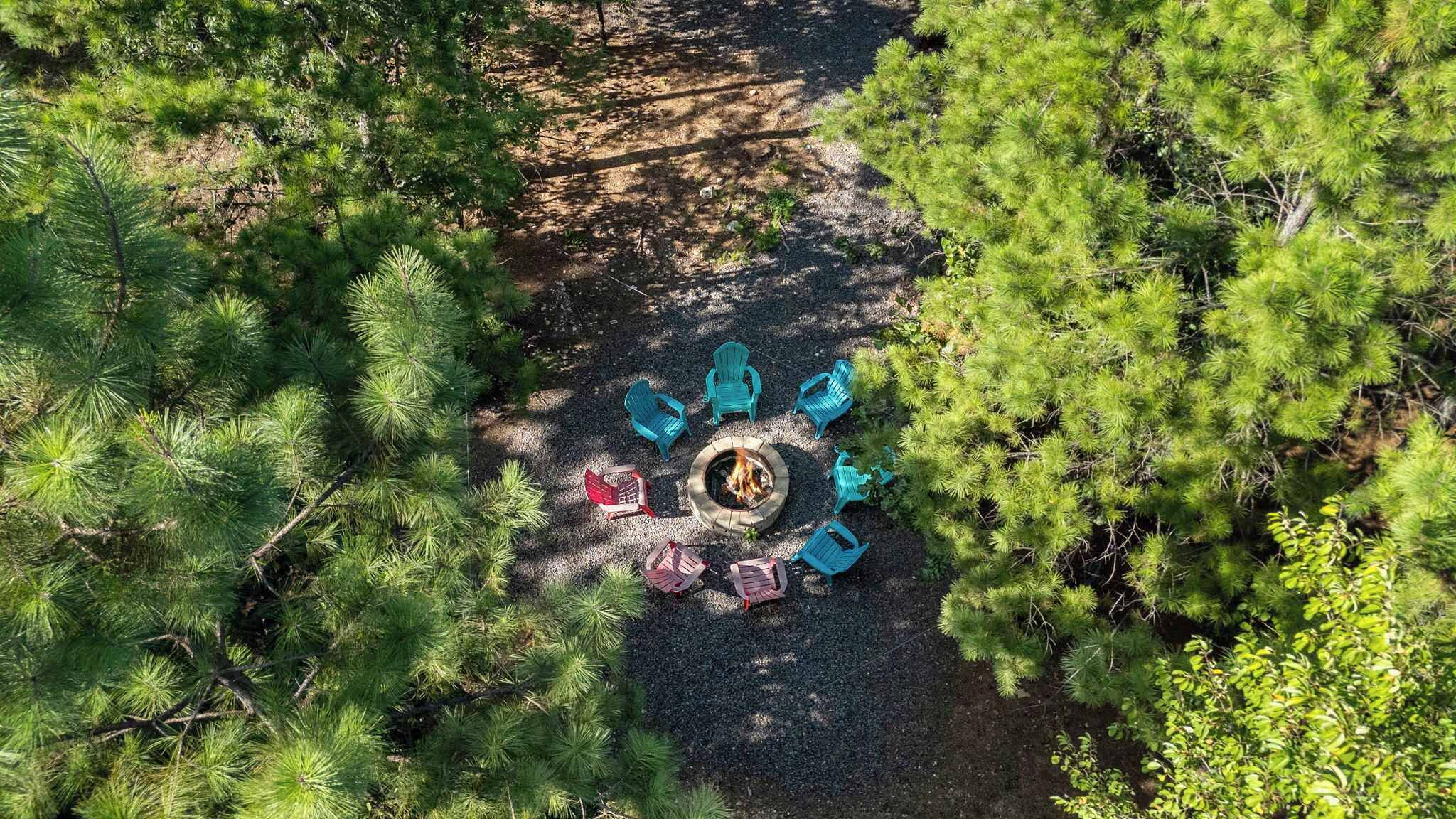 An aerial view of a campsite with a circular seating arrangement around a central fire pit, surrounded by dense pine trees.