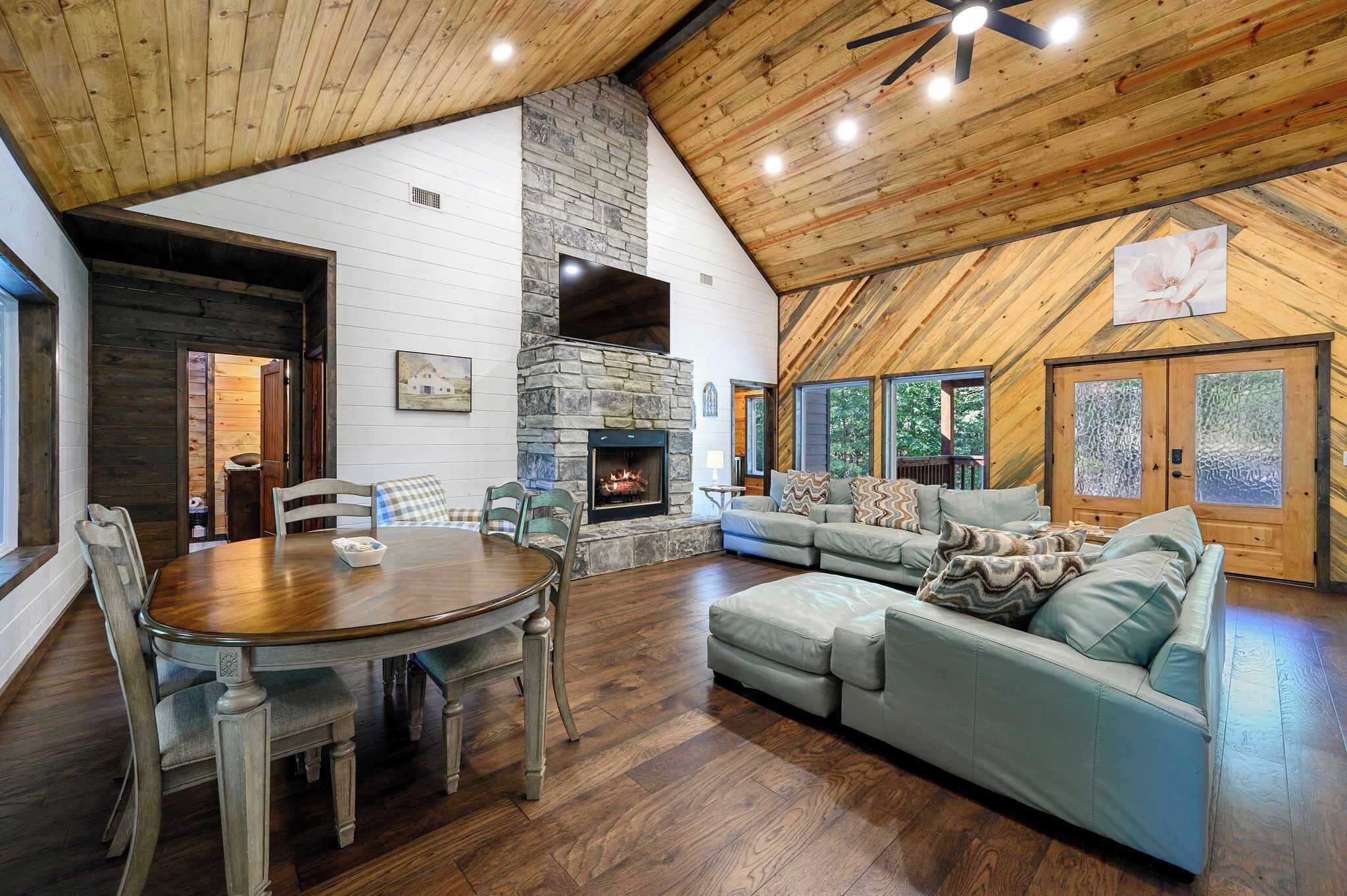 A spacious living area with a stone fireplace, wooden ceiling, and a large window overlooking greenery.