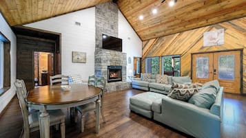 A spacious living area with a stone fireplace, wooden ceiling, and a large window overlooking greenery.