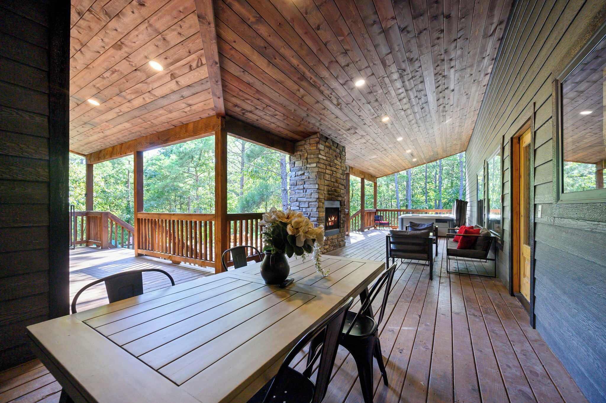 A covered outdoor dining area with a stone fireplace, wooden deck, and a view of the surrounding forest.