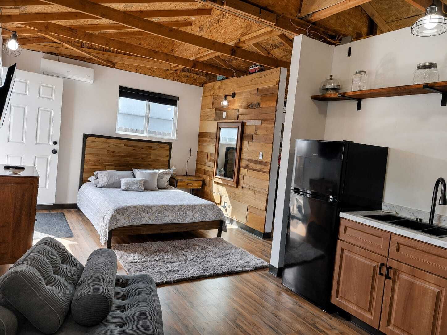 Una camera da letto con una testiera in legno, un letto con cuscini, un comodino, una mensola a muro con barattoli e un frigorifero nero.