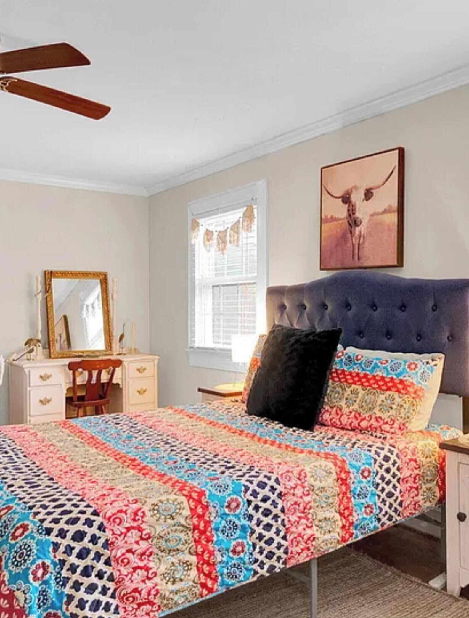 A bedroom with a tufted headboard, a patterned quilt, a mirror, and a window with shutters.