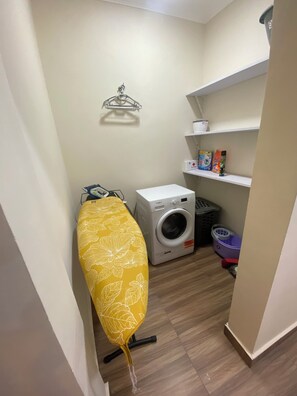 Uma lavanderia com máquina de lavar, varal, prateleiras com produtos de limpeza e um cesto de roupa.