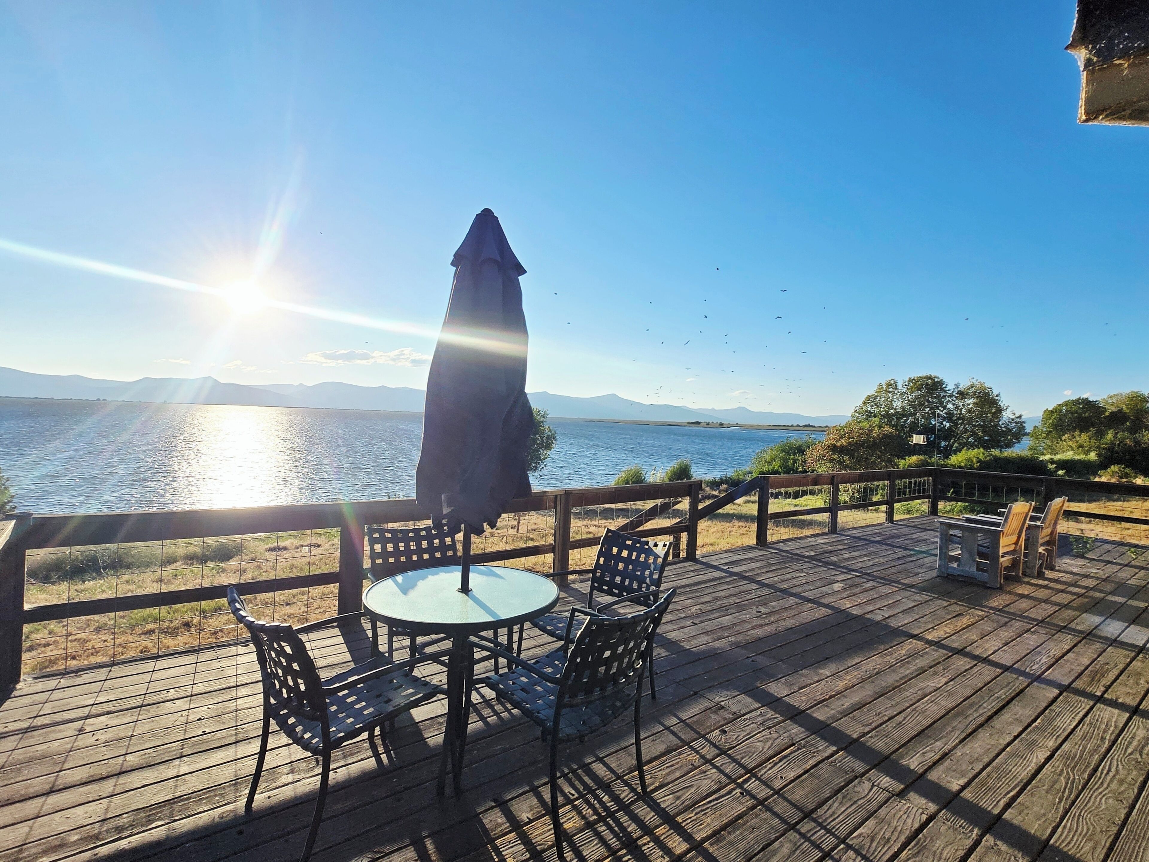 A wooden deck with a table and chairs, overlooking a body of water with mountains in the distance.