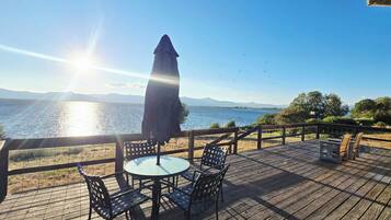 A wooden deck with a table and chairs, overlooking a body of water with mountains in the distance.