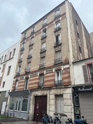 A multi-story building with a ground-floor entrance, a sign indicating a restaurant, and a row of parked scooters. - Simple, affordable studio apartment near Paris (Clichy)