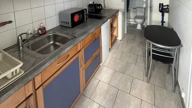 A compact kitchen with wooden cabinets, a sink, a microwave, and a small dining area with a chair. - Simple, affordable studio apartment near Paris (Clichy)
