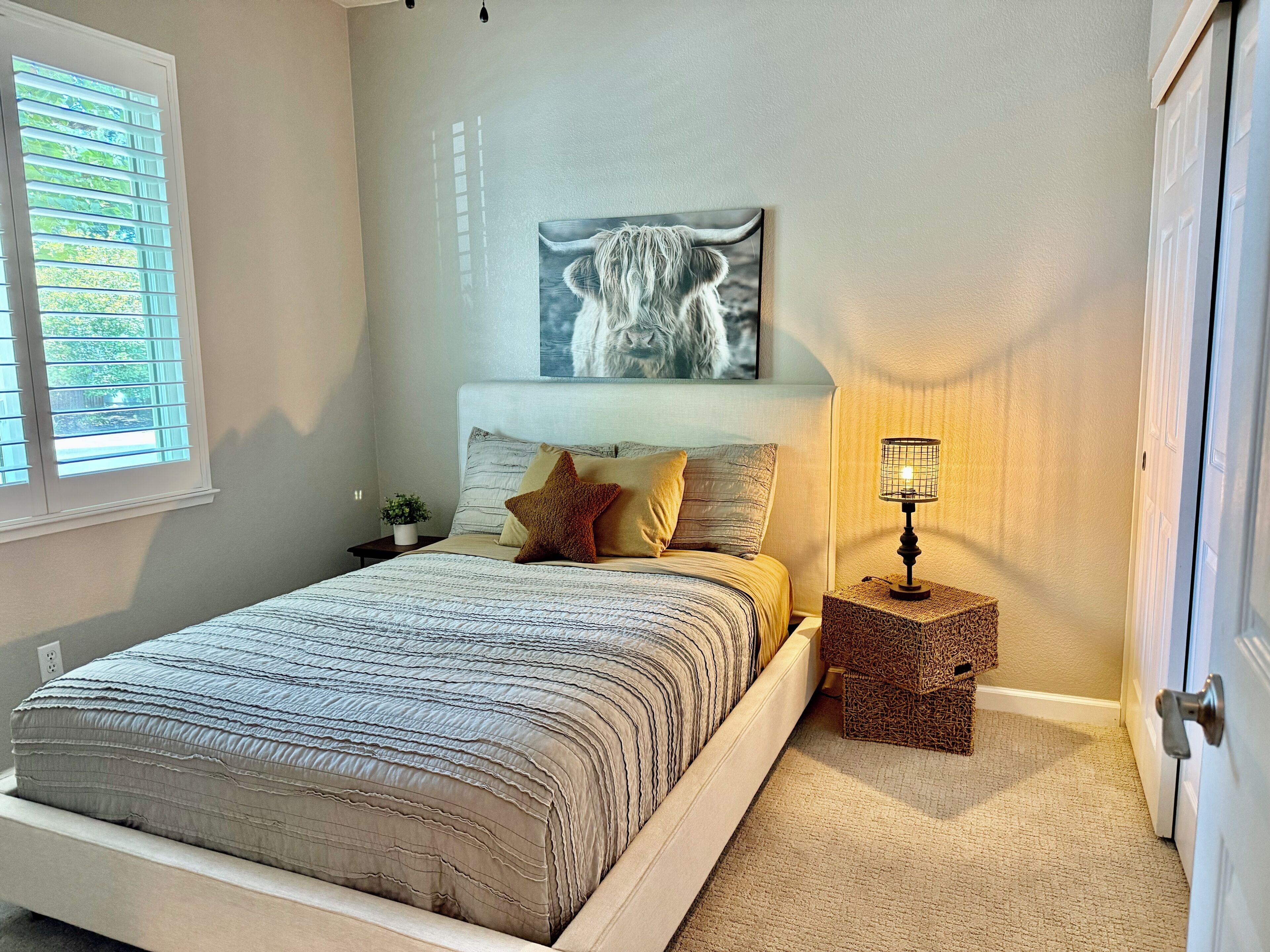 A bedroom with a bed, a nightstand, a lamp, a wall art piece, and a window with blinds.