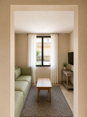 Ein Zimmer mit einem grünen Sofa, einem kleinen Holztisch, einem Fernseher auf einem Schrank und einem Fenster mit Vorhängen.