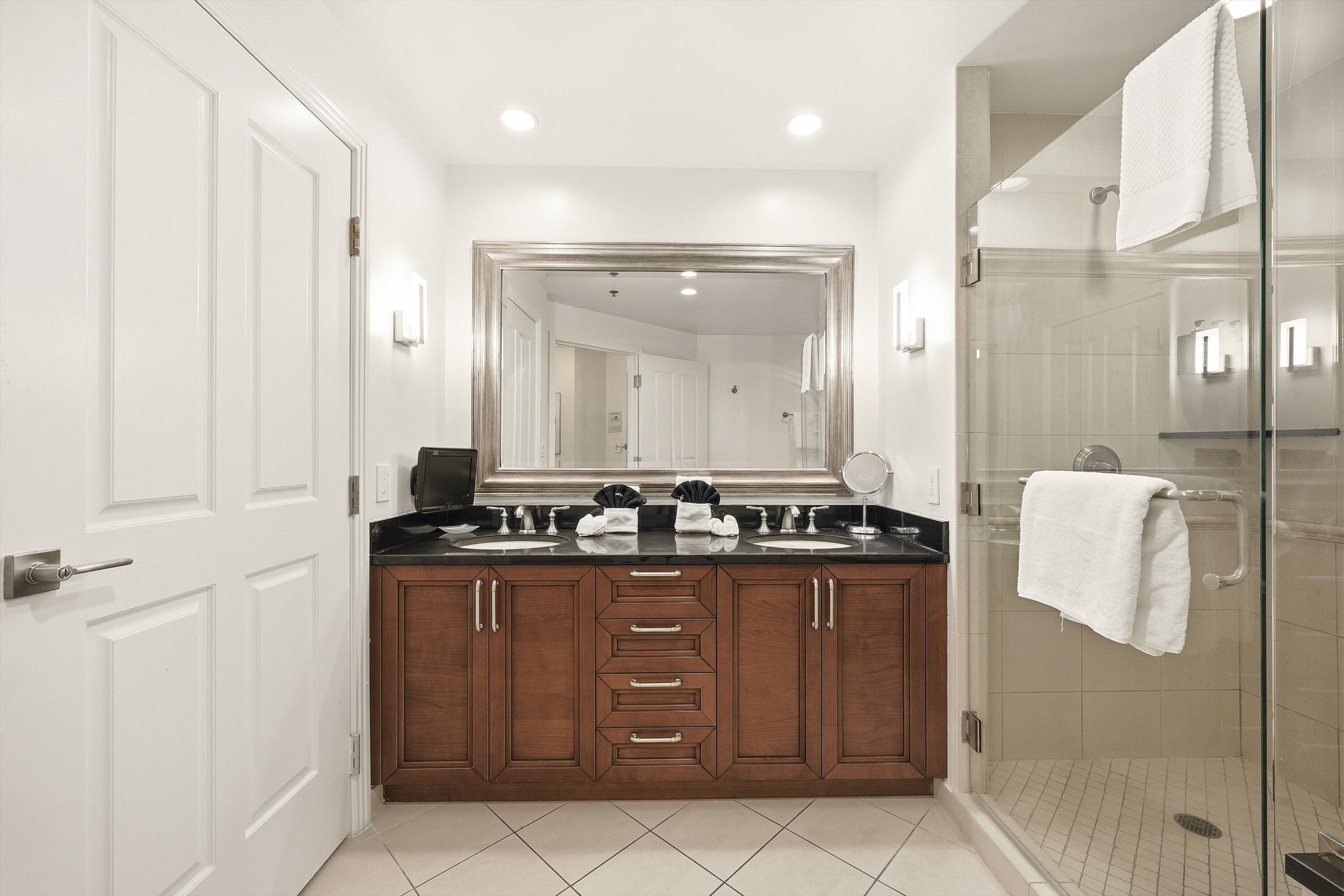 Une salle de bain avec un meuble-lavabo à deux vasques, un grand miroir, une cabine de douche en verre et une serviette blanche accrochée à la porte de la douche.