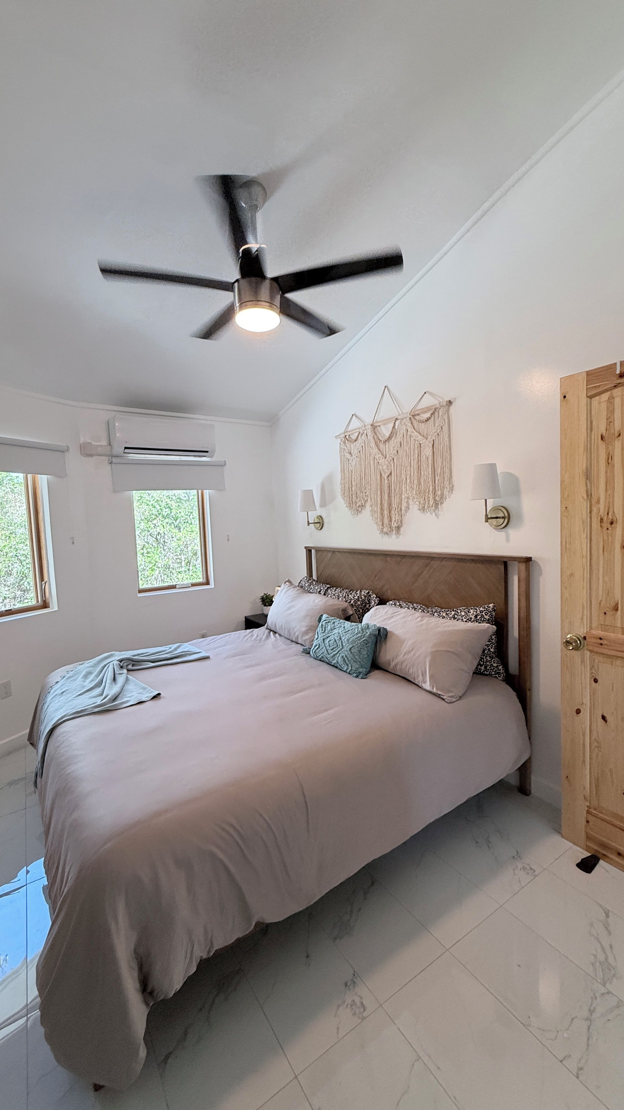 Un dormitorio con cama, ventilador de techo, unidad de aire acondicionado, puerta de madera y un adorno de macramé en la pared.