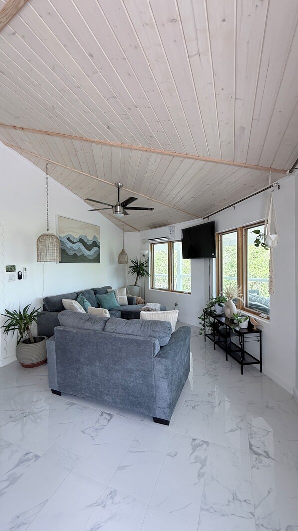 A modern living room with a grey sofa, a ceiling fan, and a TV mounted on the wall.
