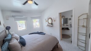 Un dormitorio con cama, ventilador de techo, ventana y baño visible a través de una puerta abierta.