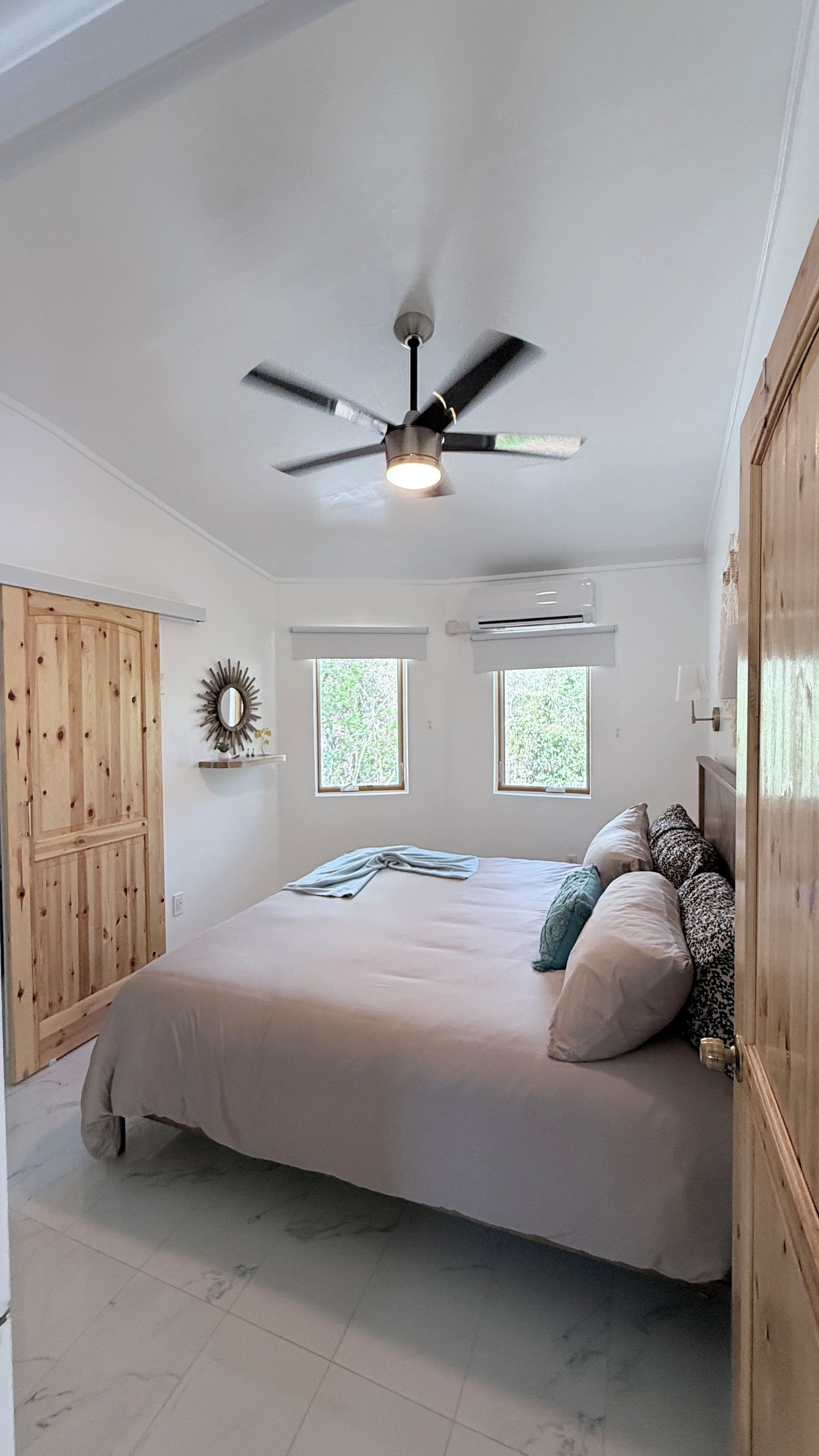 A bedroom with a bed, a ceiling fan, two windows, a wooden door, and a wall-mounted air conditioning unit.