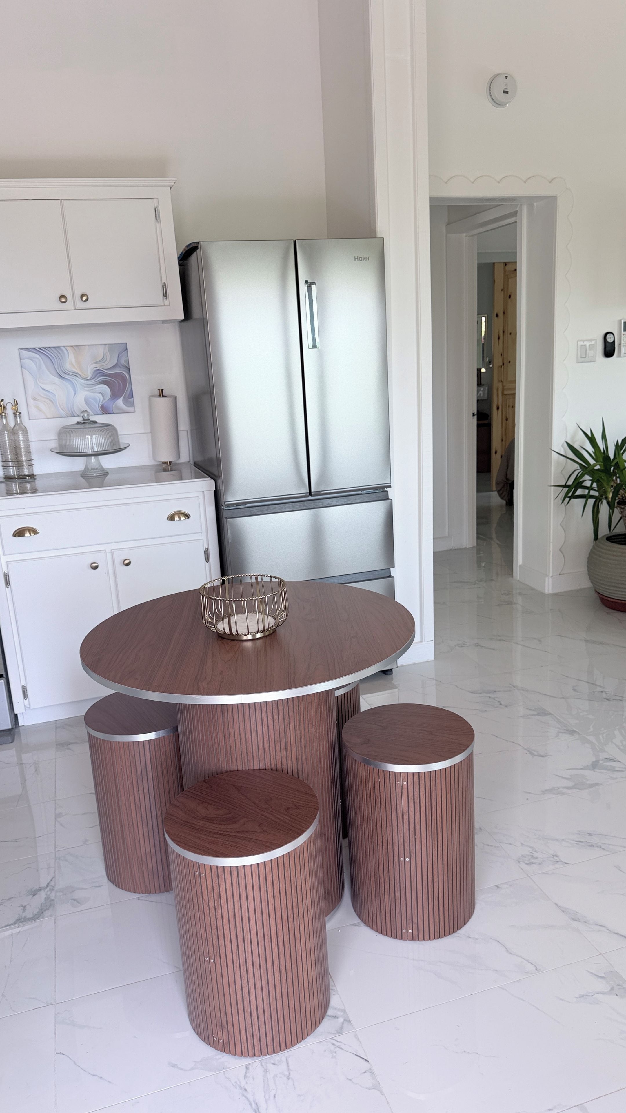 A modern kitchen with a stainless steel refrigerator, a round wooden dining table with four stools, and a marble floor.