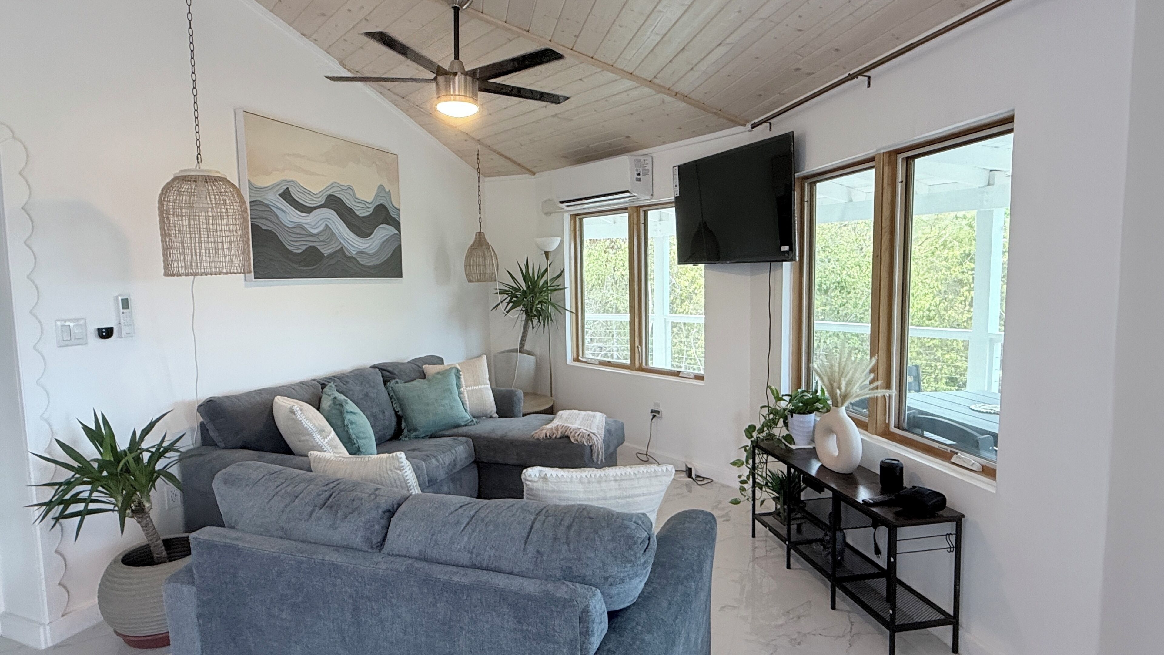 A modern living room with a sectional sofa, a ceiling fan, a flat-screen TV, and large windows.