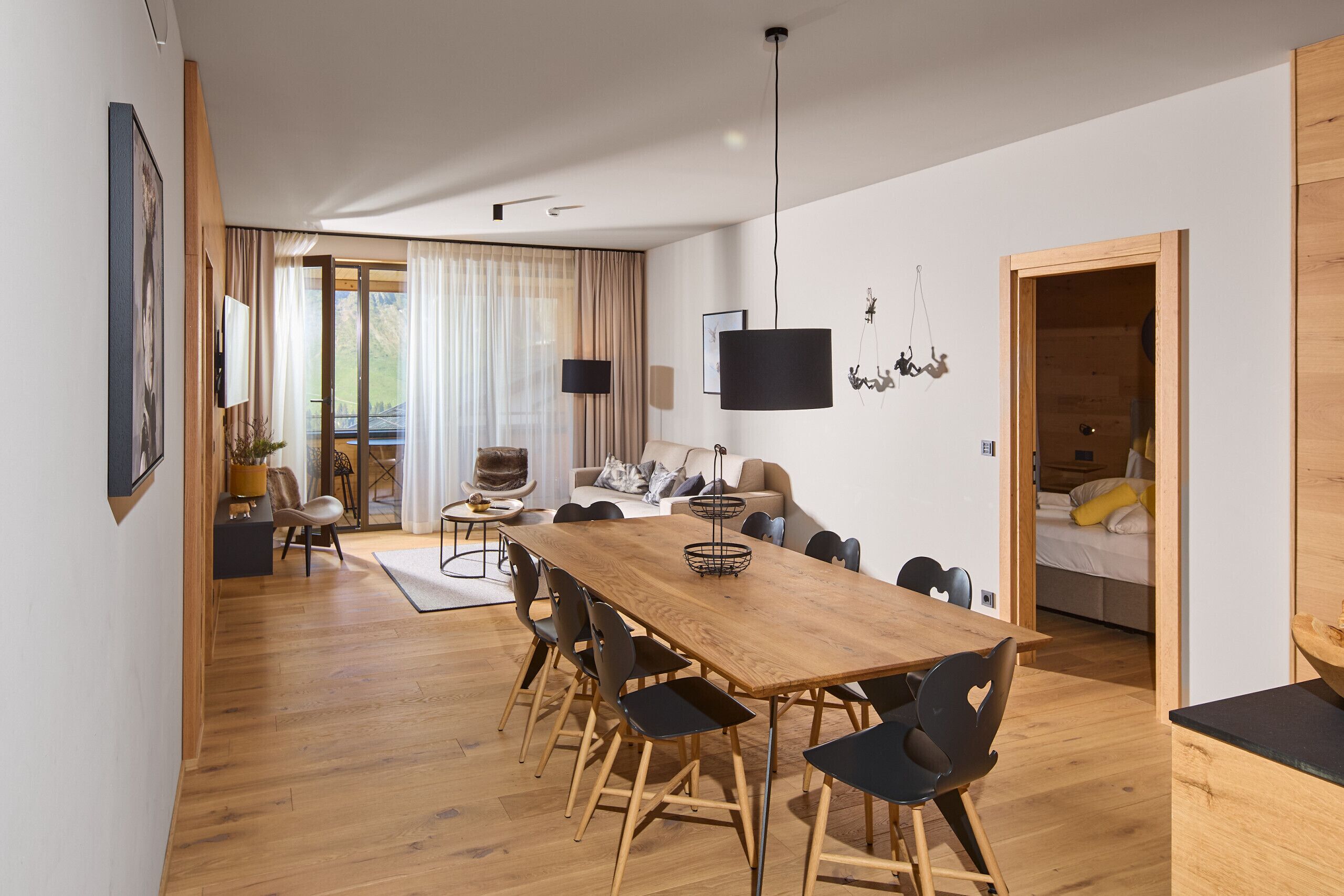 Un espacio moderno de estar con una mesa de comedor de madera, sillas negras, un sofá y un televisor montado en la pared.