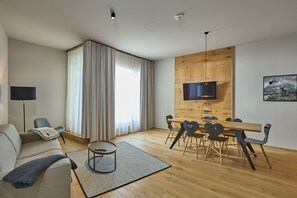 A modern living room with a sofa, a coffee table, a dining table with chairs, a TV mounted on a wooden wall, and a large window with curtains. - 8005 Alpin Retreat Grand 5 (Schröcken)