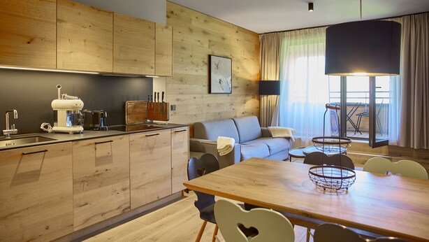 Eine moderne Küche mit Holzausstattung, ein Essbereich mit Holztisch und Stühlen, eine Couch und ein großes Fenster mit Vorhängen.