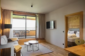 A modern hotel room with a sofa, a small table, a bed with a headboard, a television mounted on the wall, and a sliding glass door leading to an outdoor area with chairs. - 8007 Alpine Retreat 7 (Schröcken)