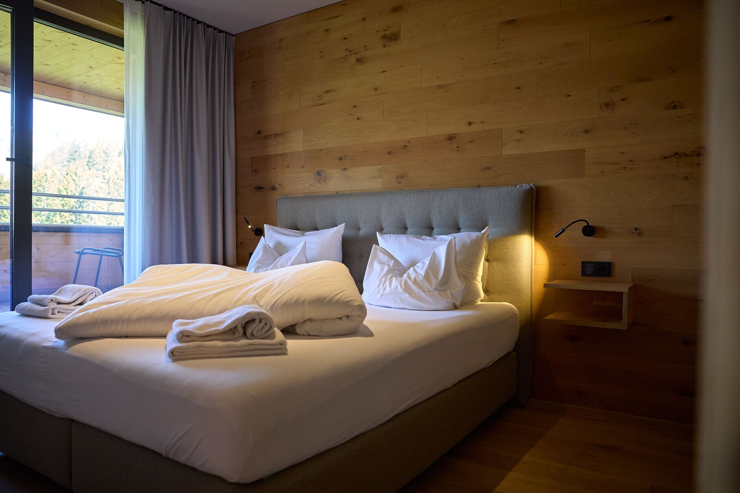 A neatly made bed with pillows and folded towels, a bedside table with a lamp, and a view of the outdoors through a window.