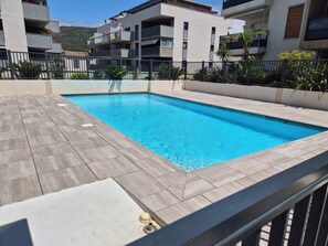 A swimming pool with a tiled deck, surrounded by a metal fence, and adjacent to a multi-story building with balconies. - Air-conditioned one-bedroom apartment near the beach with parking and a pool (Le Lavandou)