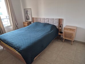 A bedroom with a bed, a bedside table, a nightstand, a wall-mounted lamp, and a window with curtains. - Air-conditioned one-bedroom apartment near the beach with parking and a pool (Le Lavandou)