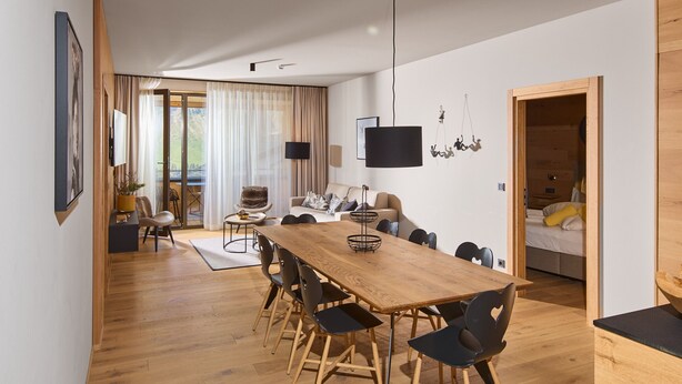 Een moderne woonruimte met een houten eettafel, zwarte stoelen, een bank en een aan de muur bevestigde televisie.