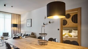 Een moderne hotelkamer met een houten eettafel, een bank, een bed met gele handdoeken en een groot raam met gordijnen.
