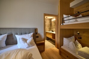 A hotel room with a bunk bed, a single bed, a bedside table, a nightstand, a mirror, a wooden cabinet, and a small bathroom with a sink and mirror. - 8013 Alpine Residence 13 (Schröcken)