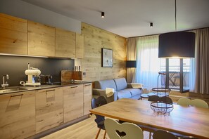 A modern kitchen with wooden cabinets, a dining area with a wooden table and chairs, a sofa, and a large window with curtains. - 8016 Alpin Retreat 16 (Schröcken)