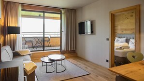 Ein modernes Hotelzimmer mit einer Couch, einem kleinen Tisch, einem Bett mit Kopfteil, einem an der Wand befestigten Fernseher und einer Schiebetür zu einem Außenbereich mit Stühlen.