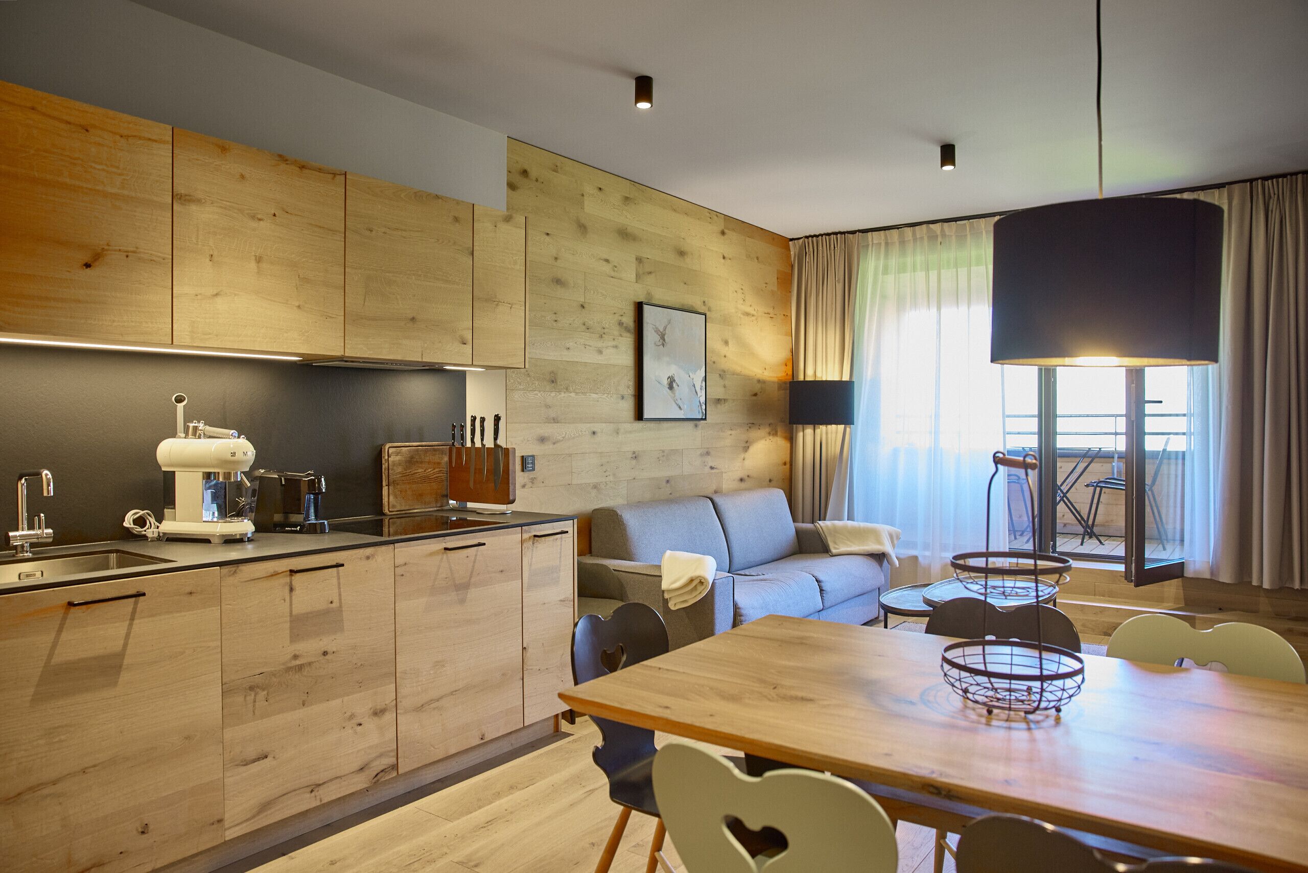 A modern kitchen with wooden cabinets, a dining area with a wooden table and chairs, a sofa, and a large window with curtains.