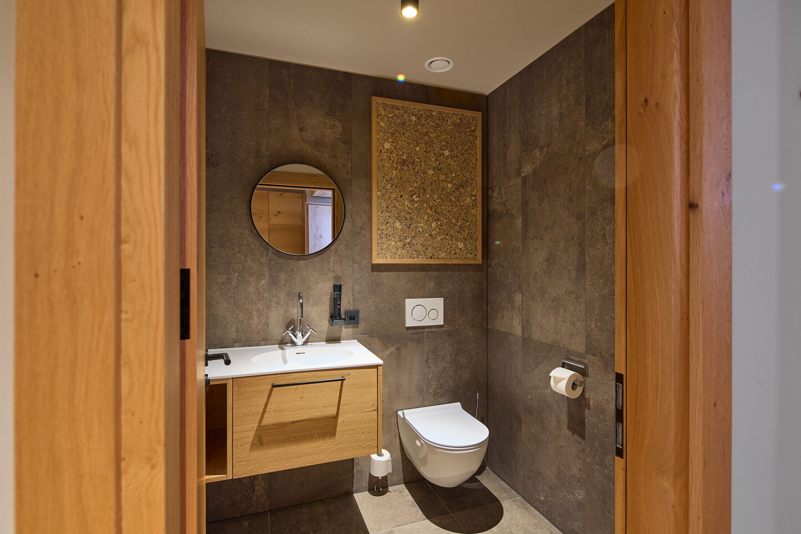 Een moderne badkamer met een houten wastafelmeubel, een ronde spiegel, toilet en een wastafel bevestigd aan de muur.