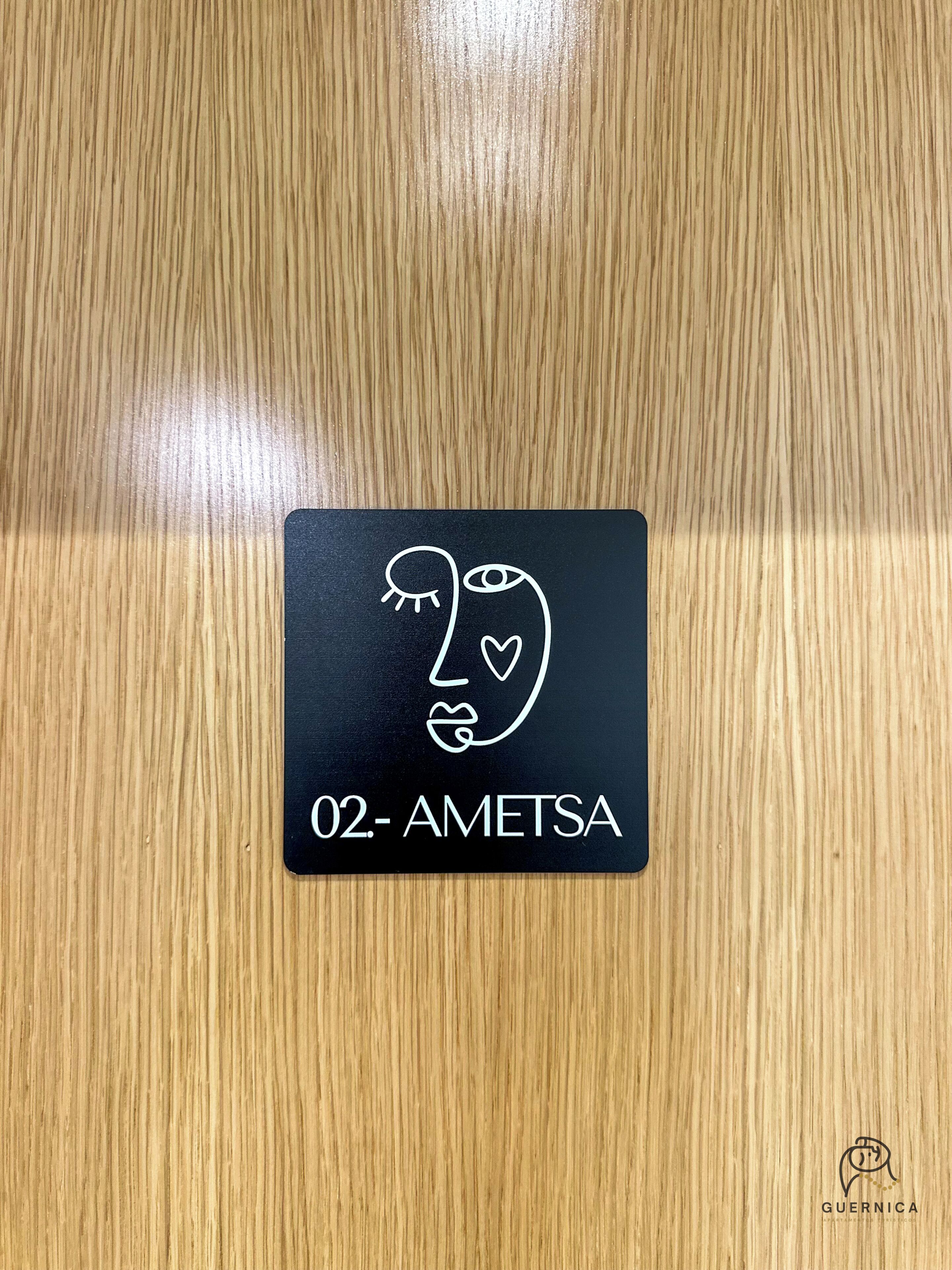 Un letrero negro con un dibujo blanco de un rostro y el texto '02- AMETSA' sobre una superficie de madera.