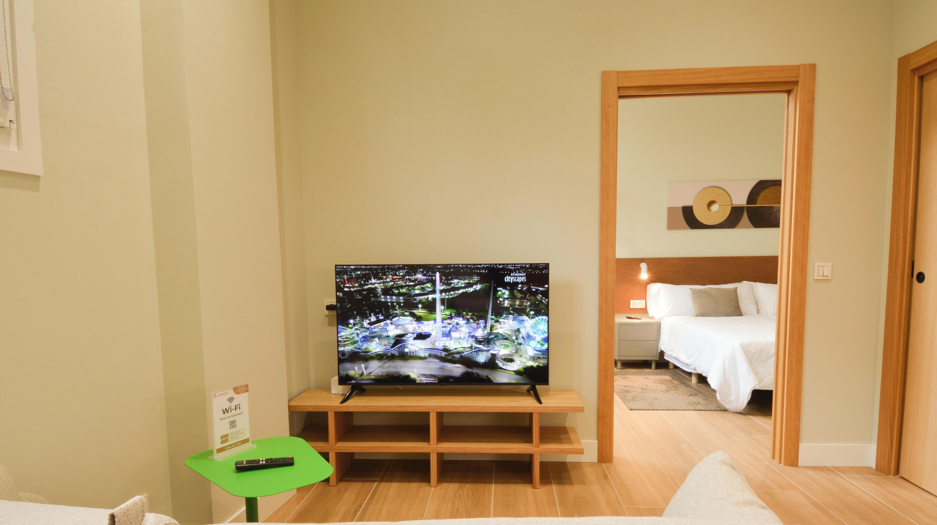 Habitación de hotel con televisor sobre un soporte de madera, cama con ropa blanca, una mesita con control remoto y un cartel que indica la disponibilidad de Wi-Fi.
