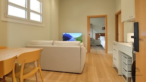 A modern living room with a sofa, a dining table, and chairs. There is a television mounted on the wall. - GUERNICA Suites Downtown (Gernika-Lumo)