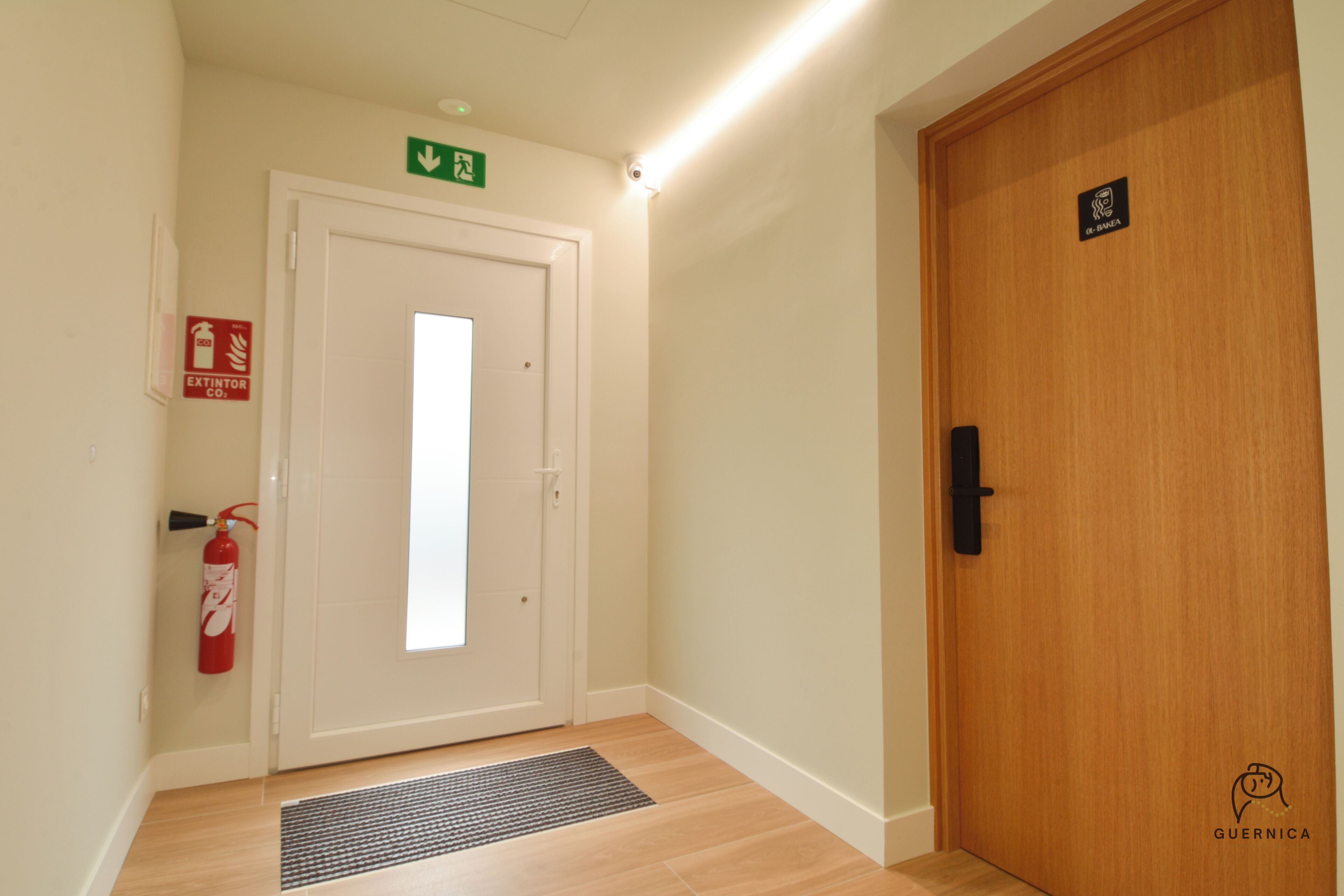 Un pasillo con una puerta blanca, un extintor, un letrero de salida de emergencia verde y una puerta de madera con picaporte negro.