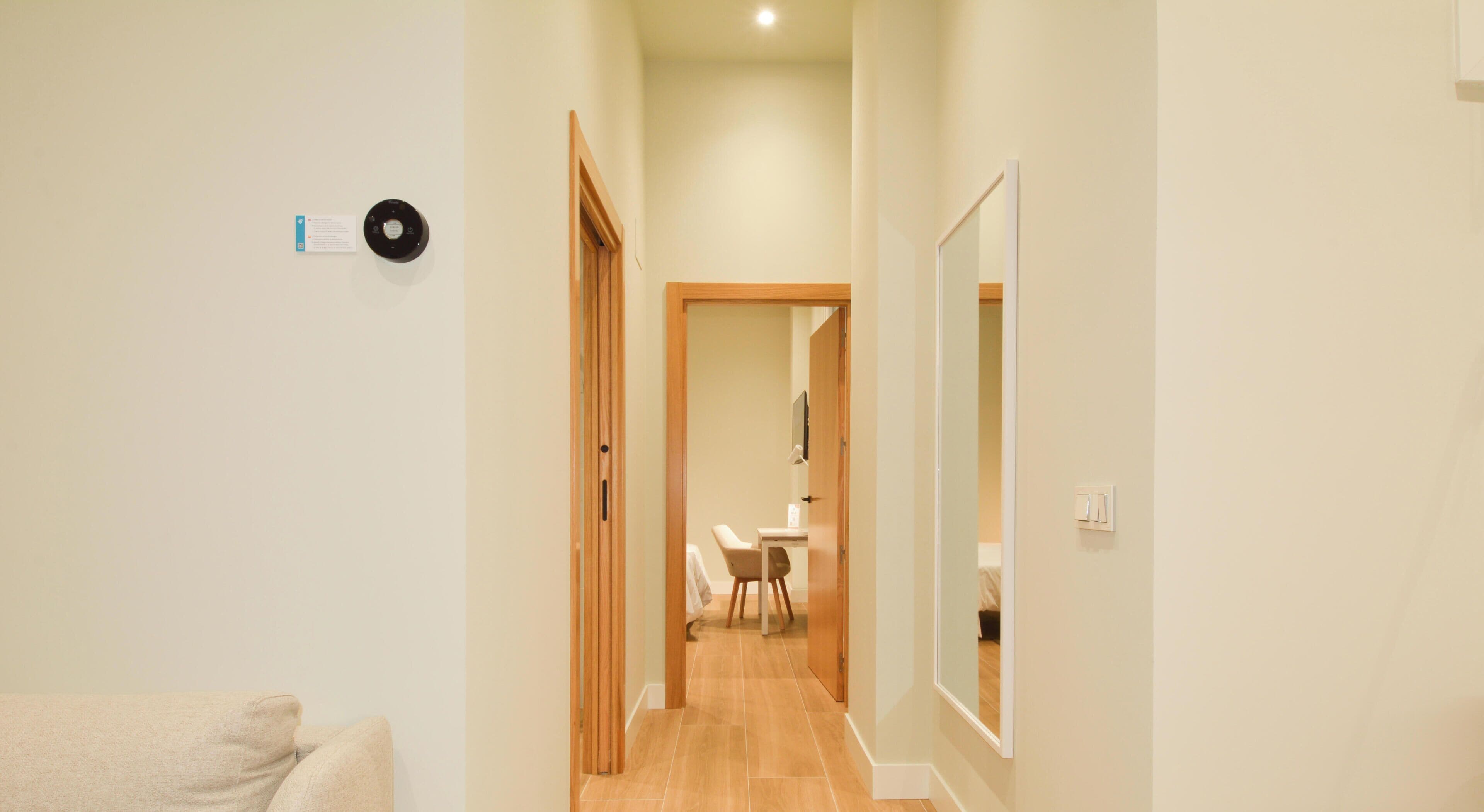 A hallway with a wooden door leading to a room with a bed and a desk.