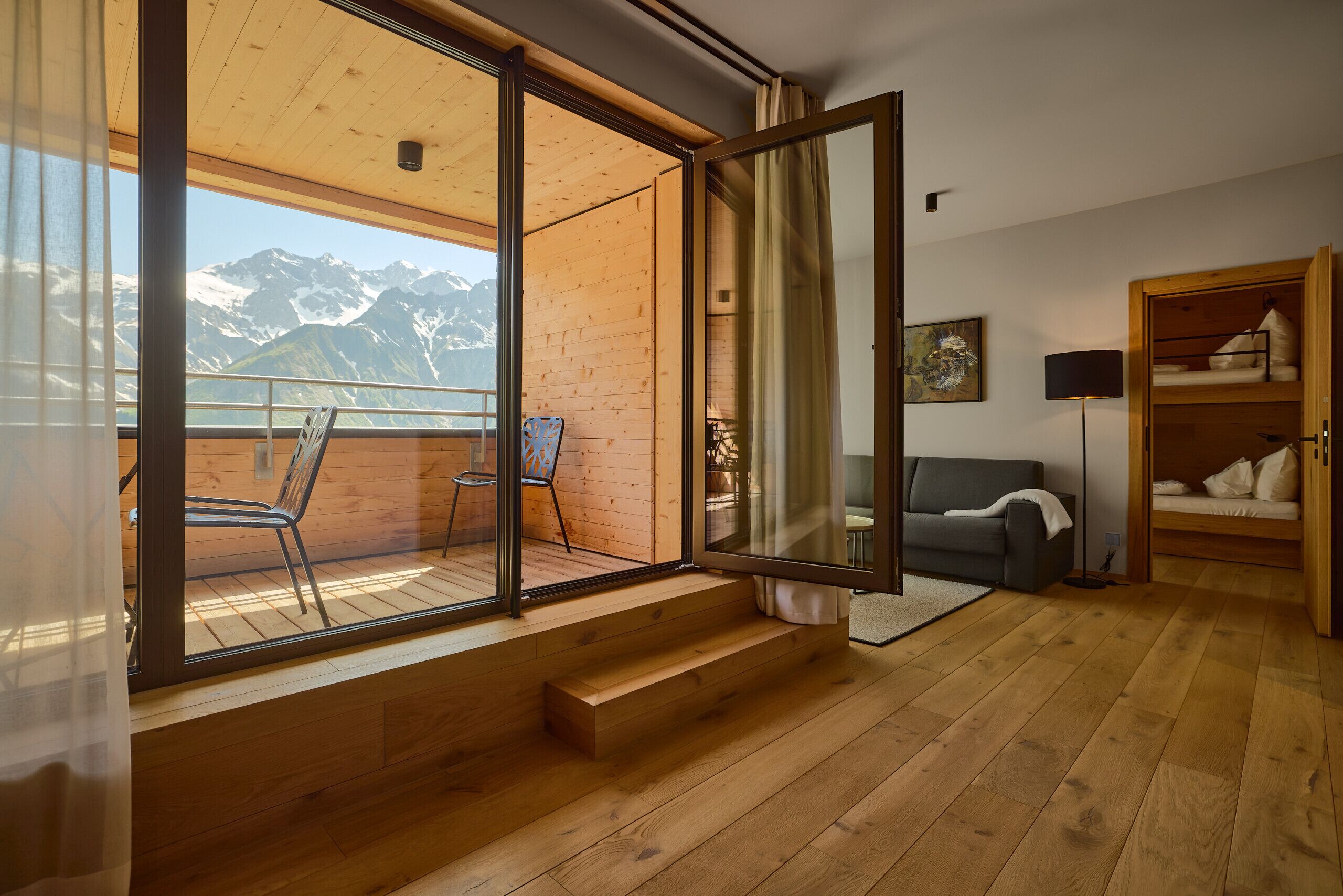 A room with a wooden floor, a sofa, a chair, a bed, and a window with a mountain view.