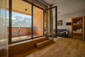 A room with a wooden floor, a sofa, a chair, a bed, and a window with a mountain view. - 8018 Summit Residence 18 (Schröcken)
